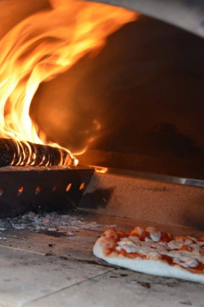 Wood-Burning Pizza Oven Cooking – Florida Outdoor Kitchen - Paradise Outdoor Kitchens A pizza cooking inside a wood-burning oven with flames rising inside a modern outdoor kitchen setup.